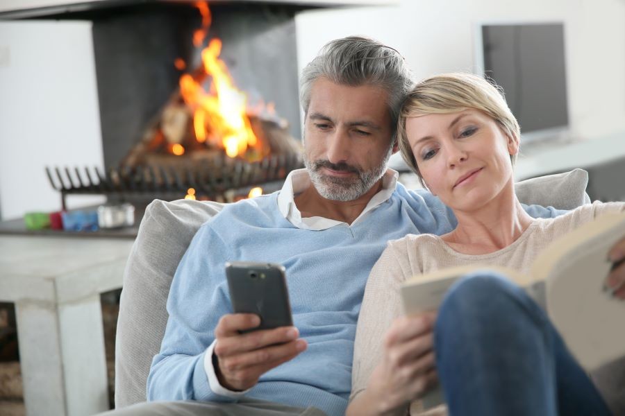 Two people sitting next to each other on a couch in front of a fireplace, looking at a smartphone and reading a book.
